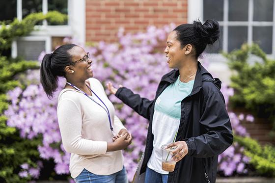 Photo of two women stand together laughing outside of a redbrick Chatham University academic building